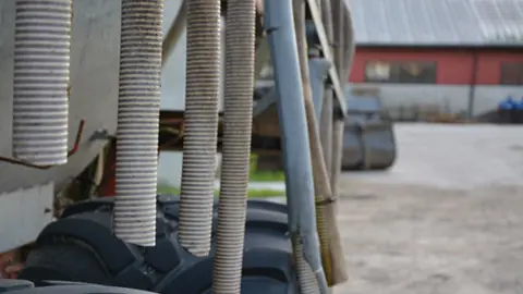 Slangar som hänger ner från en jordbruksmaskin. Maskinen står på gårdsplan på ett lantbruk.